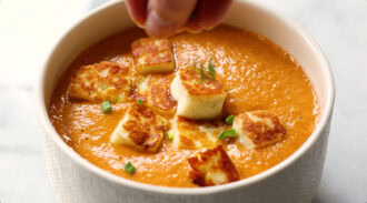A hand sprinkles herbs onto a bowl of creamy orange soup topped with golden-brown cheese cubes.