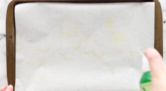 A hand sprays cooking oil onto parchment paper lined on a baking sheet.