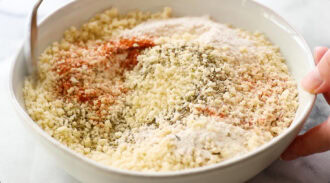 A hand holds a bowl filled with breadcrumbs, flour, and various spices, being mixed with a spoon.