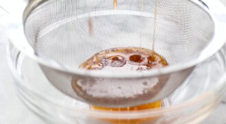 A mesh strainer filters a golden brown liquid into a glass bowl below.