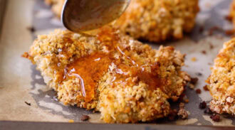 A spoon drizzles honey over a piece of baked breaded chicken on a parchment-lined baking sheet.