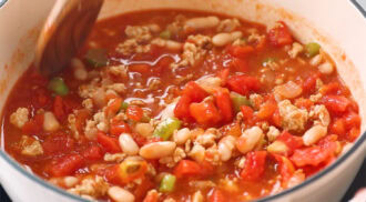 A close-up of a pot of chili with ground meat, diced tomatoes, white beans, onions, and green peppers being stirred with a wooden spoon.