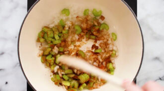Diced onions, celery, and spices are being stirred with a wooden spoon in a white pot on a marble countertop.