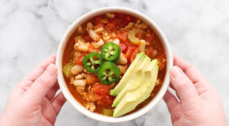 A bowl of chili with white beans, ground meat, tomatoes, celery, sliced avocado, and sliced jalapeño, held in two hands over a marble surface.