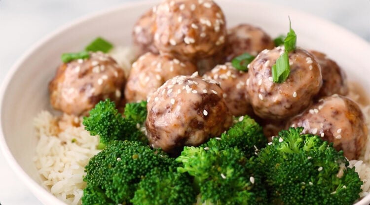 Bowl of white rice topped with glazed meatballs, sprinkled with sesame seeds and green onions, served with steamed broccoli.