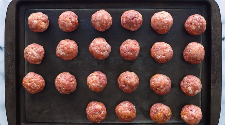 Twenty raw meatballs arranged in four rows on a dark metal baking sheet, ready to be cooked.