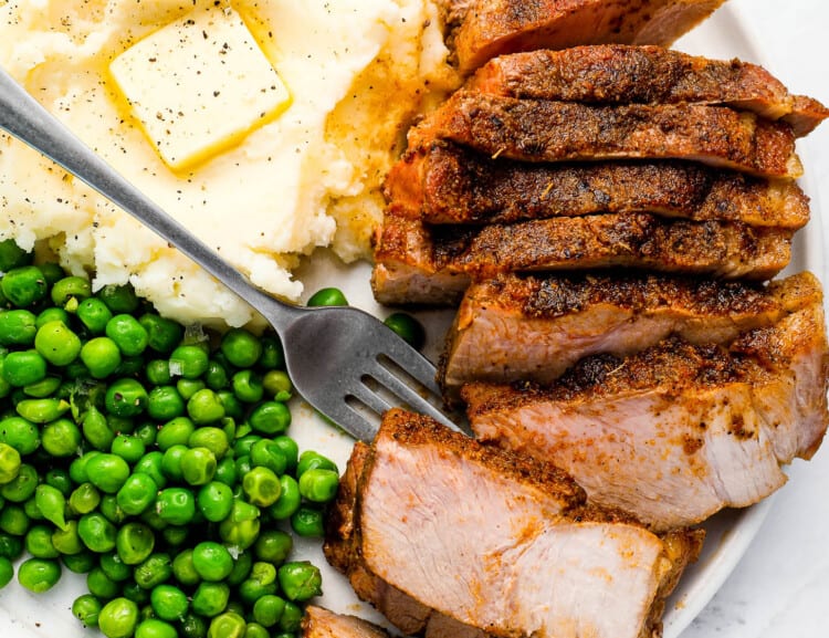 An oven baked pork chop on a dinner plate with mashed potatoes and peas.