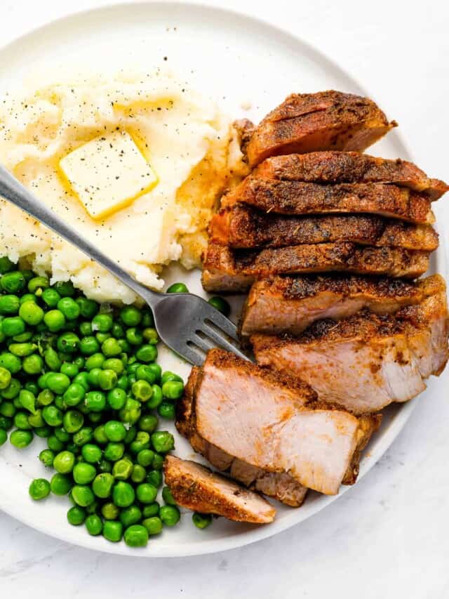 An oven baked pork chop on a dinner plate with mashed potatoes and peas.