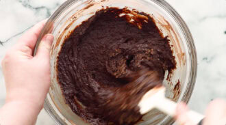 A person mixes thick chocolate batter in a glass bowl with a white spatula on a marble surface.