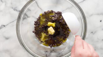 A hand pours oil into a glass bowl containing chocolate chips and a small piece of butter on a marble countertop.
