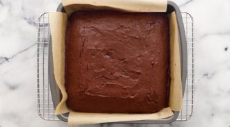 A square chocolate cake baked in a parchment-lined pan sits on a cooling rack over a marble surface.