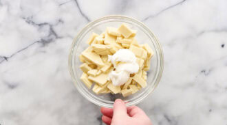 A hand holds a glass bowl filled with chopped white chocolate and a dollop of shortening on a marble countertop.