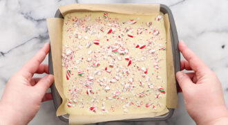 Hands holding a parchment-lined baking pan filled with white chocolate bark topped with crushed peppermint candy on a marble surface.