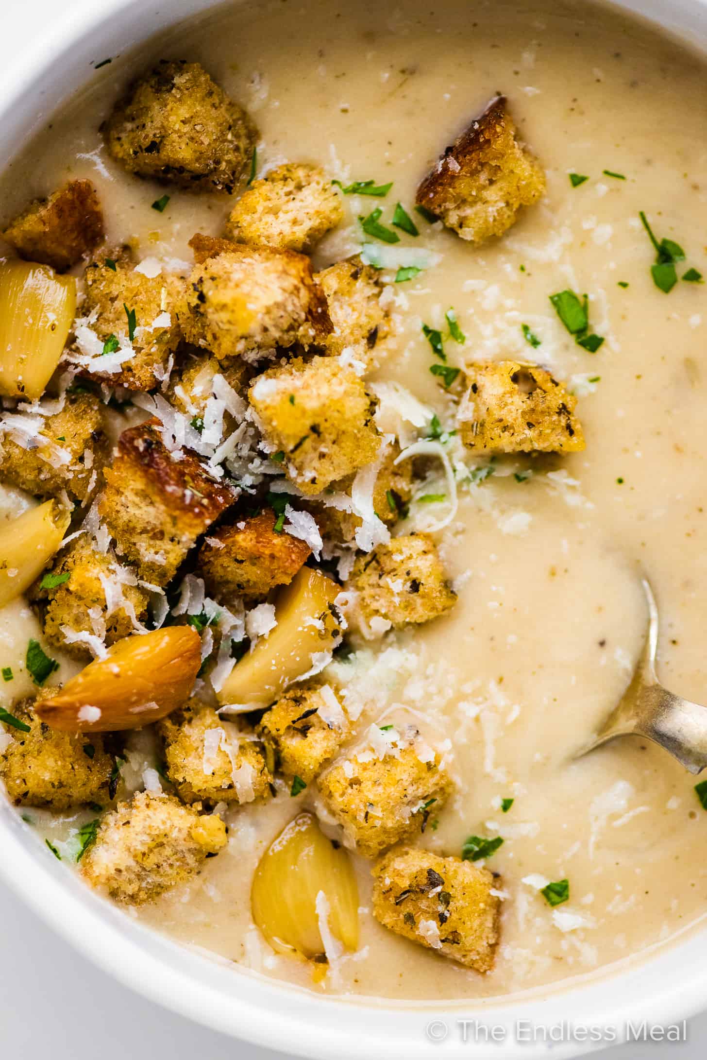 a close up of garlic soup in a white bowl topped with croutons and roasted garlic.