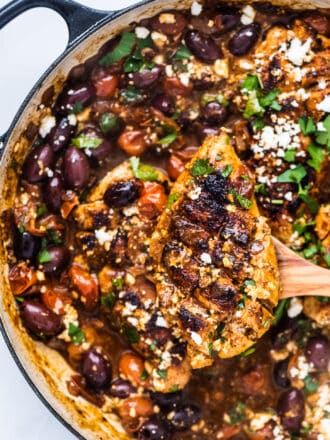 A wooden spoon lifting a piece of Greek chicken in a tomato feta sauce out of a skillet.