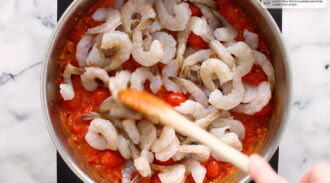 Raw shrimp are being stirred into a pot of tomato sauce with a wooden spoon on a stovetop.