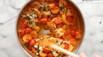 Shrimp, cherry tomatoes, herbs, and cheese cook in a stainless steel pan on a marble countertop; a hand stirs with a wooden spoon.