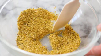 A hand uses a white spatula to mix a blend of ground spices and herbs—perfect for sprinkling over parmesan croutons—in a clear glass bowl on a marble surface.