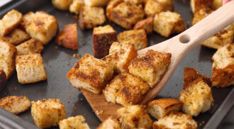 A wooden spatula lifts seasoned, baked parmesan croutons from a dark baking sheet; other croutons are scattered on the sheet.