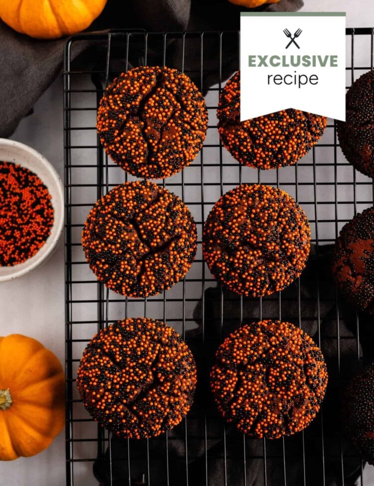 Six chocolate cookies with orange and black sprinkles on a cooling rack, surrounded by small pumpkins, a bowl of sprinkles, and a refreshing glass of blackberry lemonade.