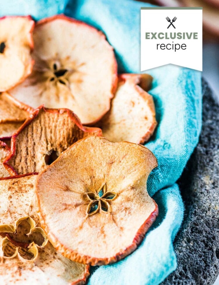 A close-up of dried apple slices with cinnamon on a teal cloth, next to a refreshing glass of blackberry lemonade.