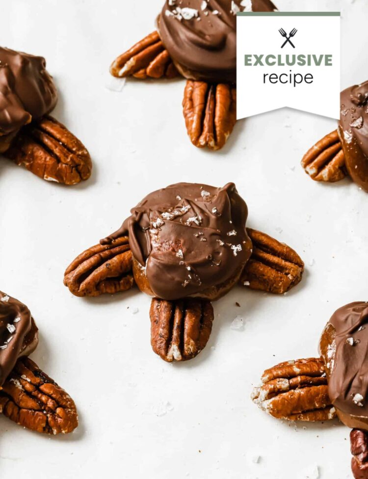 Chocolate-covered pecan clusters arranged on a white surface, topped with flaky sea salt and paired perfectly with a refreshing glass of Blackberry Lemonade, resembling turtle shapes.