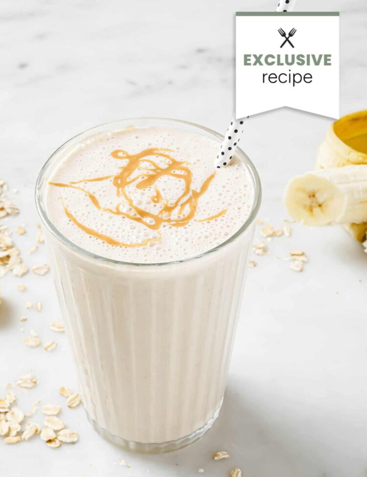 Glass of banana oat smoothie with a swirl of peanut butter on top, a polka dot straw, bananas, and oats on a marble surface—perfect alongside a refreshing Blackberry Lemonade.