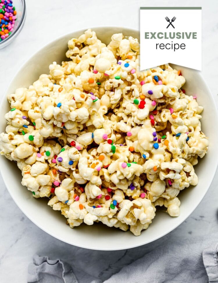 A white bowl filled with popcorn coated in a white glaze and topped with colorful round sprinkles, inspired by the vibrant hues of Blackberry Lemonade, placed on a light surface.
