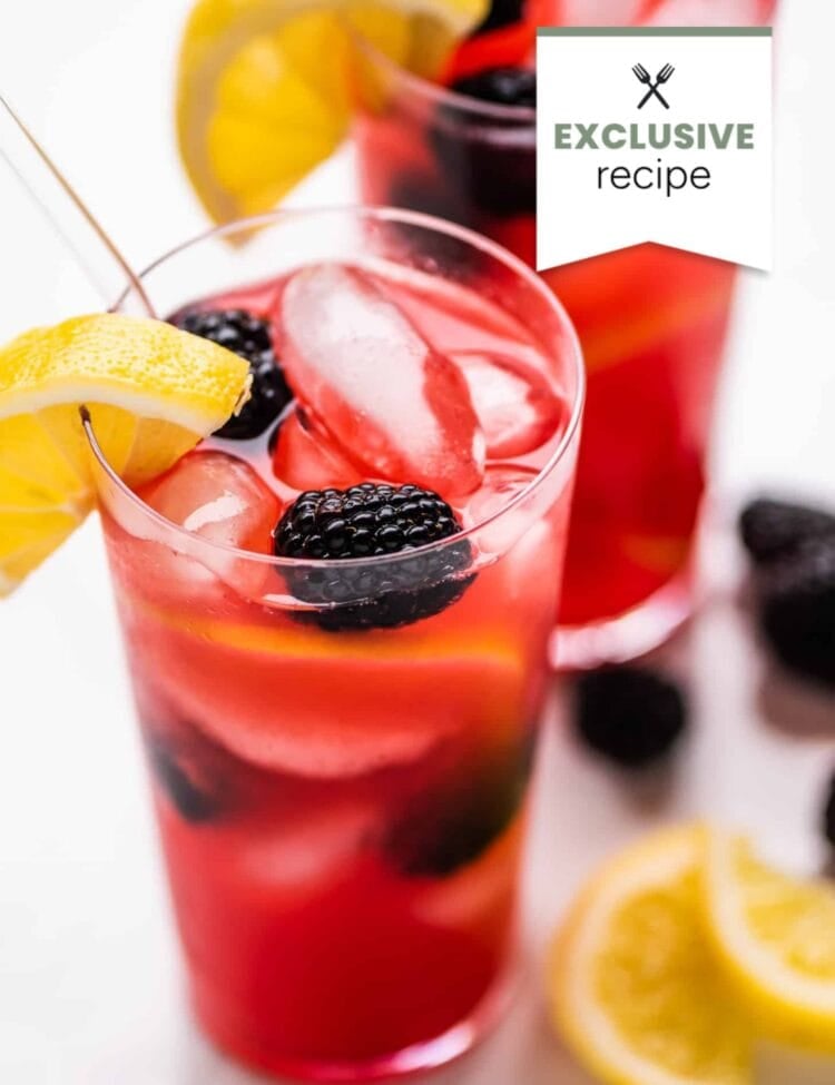 A glass of refreshing Blackberry Lemonade garnished with lemon slices and blackberries, with a second similar drink and loose fruit in the background.