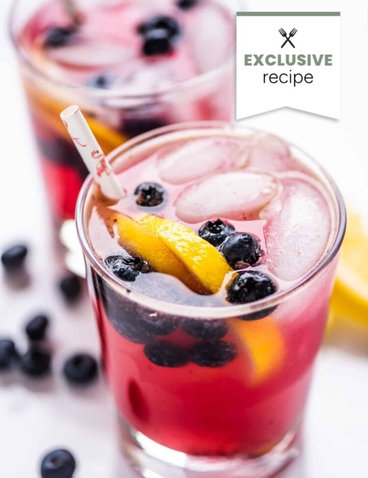 Two glasses of blackberry lemonade, a pink iced drink garnished with blueberries and lemon slices, with straws. Loose blueberries are scattered on a white surface. A label reads Exclusive recipe.