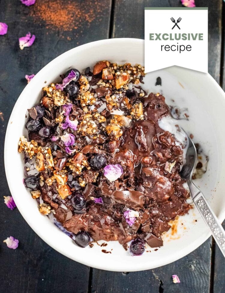 A white bowl filled with yogurt, granola, blueberries, chocolate pieces, and edible flower petals sits on a dark wooden surface with a spoon inside—perfect alongside a refreshing glass of Blackberry Lemonade.