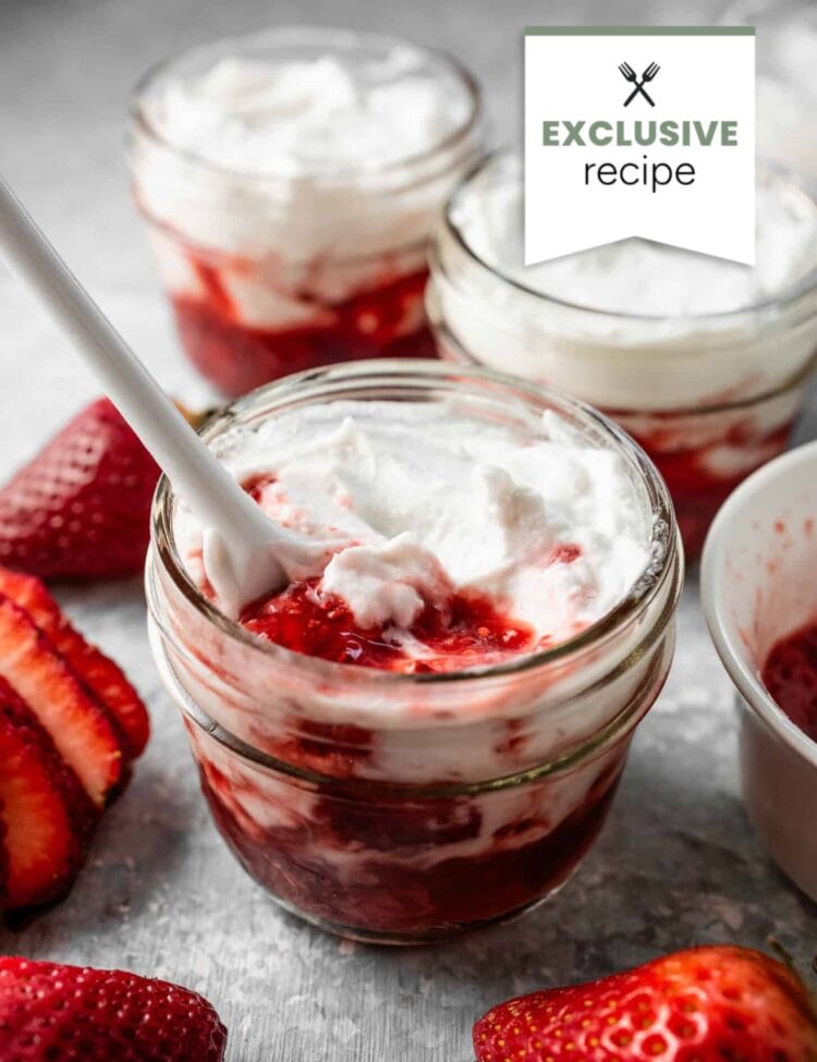 Glass jars filled with layers of strawberry sauce and whipped cream, surrounded by fresh strawberries and a hint of blackberry lemonade, with a spoon in one jar.