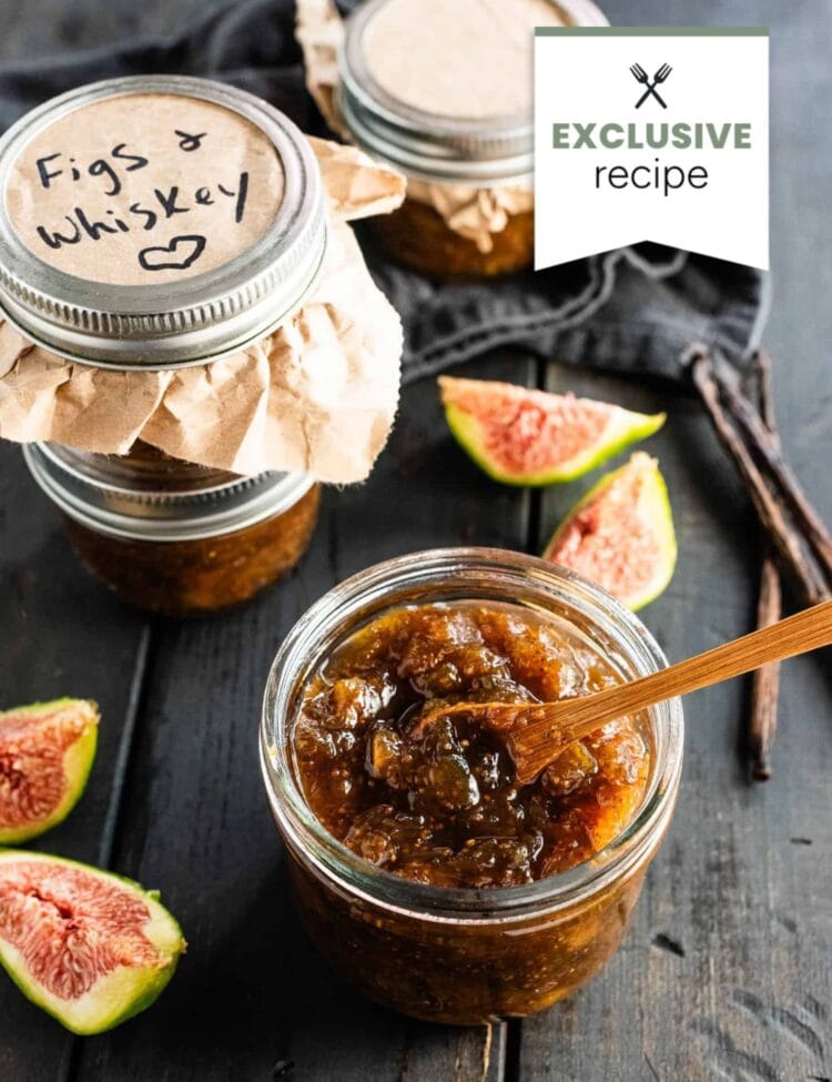 An open jar of fig and whiskey jam with a wooden spoon, surrounded by fresh fig slices, sealed jars, and a hint of blackberry lemonade on a dark surface.