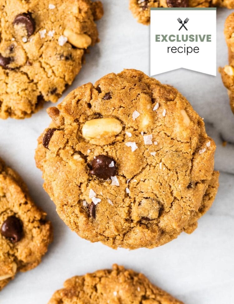 Close-up of several peanut butter cookies with chocolate chips and peanuts on a white surface, sprinkled with sea salt. A banner in the image reads “Exclusive recipe.” Enjoy with a refreshing glass of blackberry lemonade.