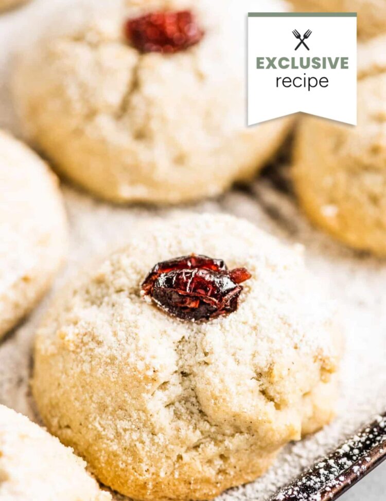 Close-up of a powdered sugar-dusted cookie topped with a dried cranberry, evoking the sweet-tart vibes of blackberry lemonade, with similar cookies in the background and an “Exclusive recipe” label in the corner.
