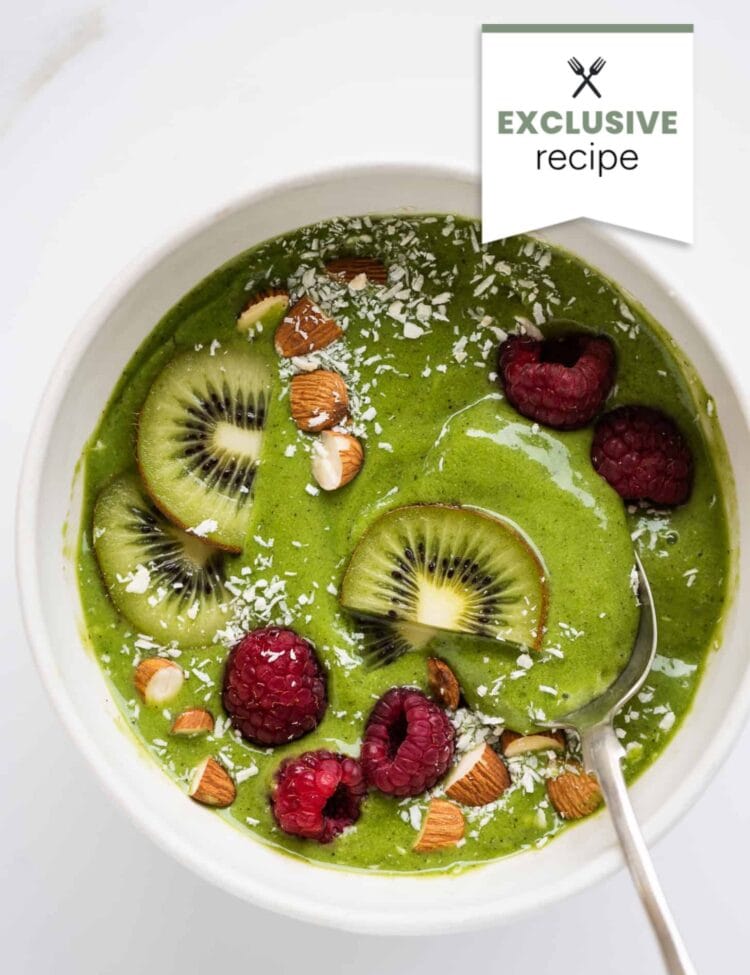 A bowl of green smoothie topped with kiwi slices, raspberries, chopped almonds, and shredded coconut, with a hint of Blackberry Lemonade flavor and a spoon placed inside.