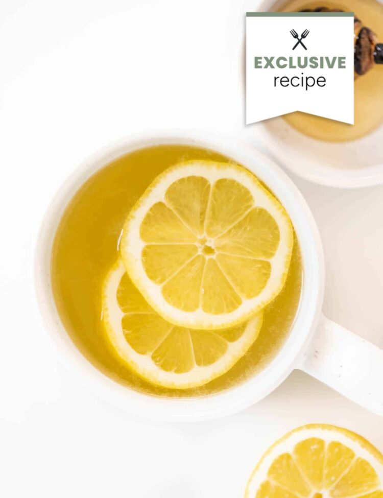 A white cup filled with hot lemon tea topped with lemon slices, alongside a cup of honey and a slice of lemon on a white surface, evokes the refreshing zest of blackberry lemonade.