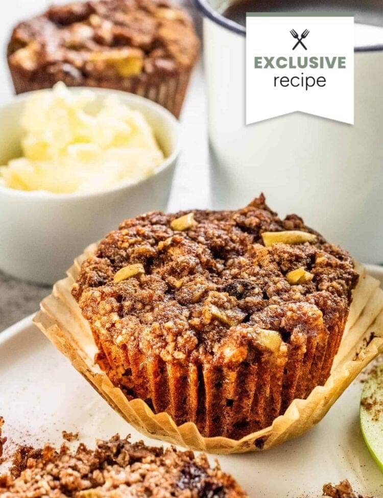 A close-up of a muffin with crumb topping on a plate, alongside a cup of coffee, a bowl of butter, and a refreshing glass of blackberry lemonade in the background.