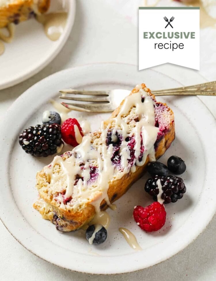 A slice of berry loaf cake with white icing on a white plate, served with assorted fresh berries and a refreshing glass of blackberry lemonade, alongside a fork.