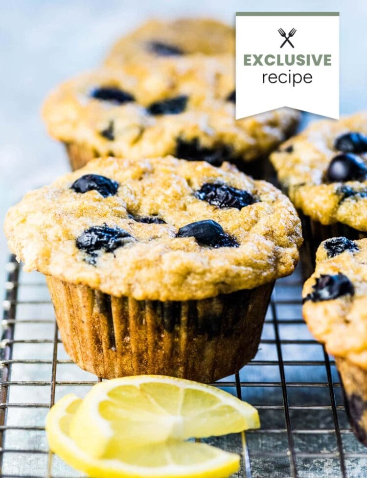 Close-up of blueberry muffins in paper liners on a cooling rack, with lemon slices in the foreground and an exclusive Blackberry Lemonade recipe label in the corner.