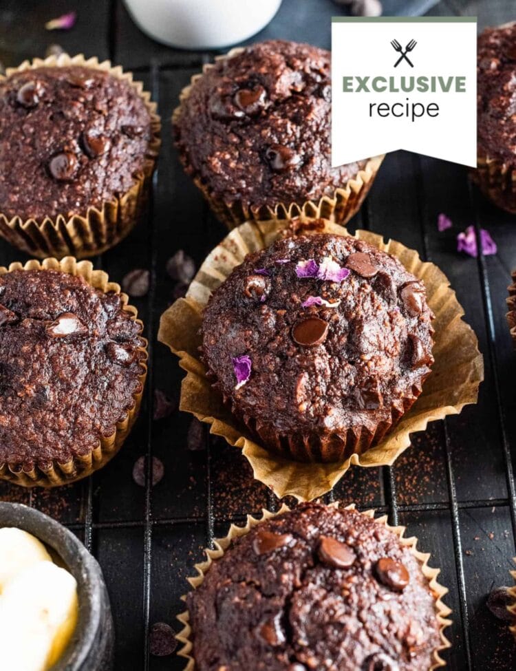 Chocolate muffins with chocolate chips are arranged on a cooling rack, some in brown paper liners, alongside a bowl of chocolate chips and a pat of butter—perfect paired with a refreshing glass of Blackberry Lemonade.