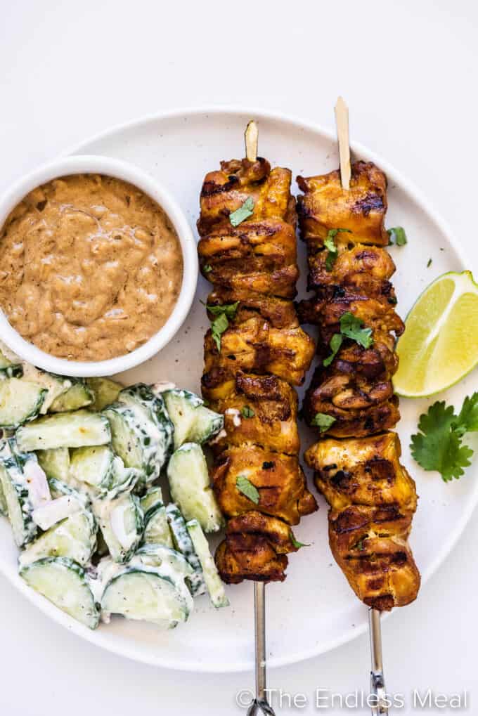 Thai Chicken Skewers on a plate next to peanut satay sauce and cucumber salad