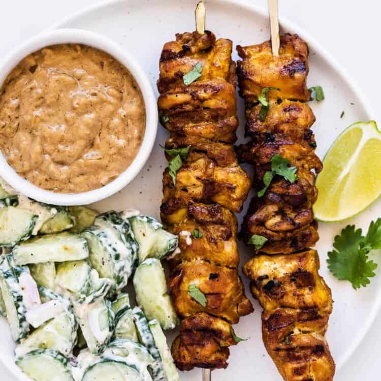 Thai Chicken Skewers on a plate next to peanut satay sauce and cucumber salad