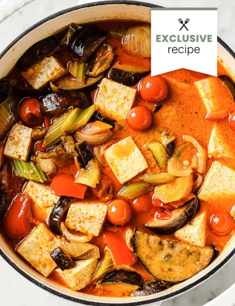 A pot filled with a colorful Thai Vegan Curry stew, including tofu, cherry tomatoes, eggplant, celery, onion, and a vibrant red broth.