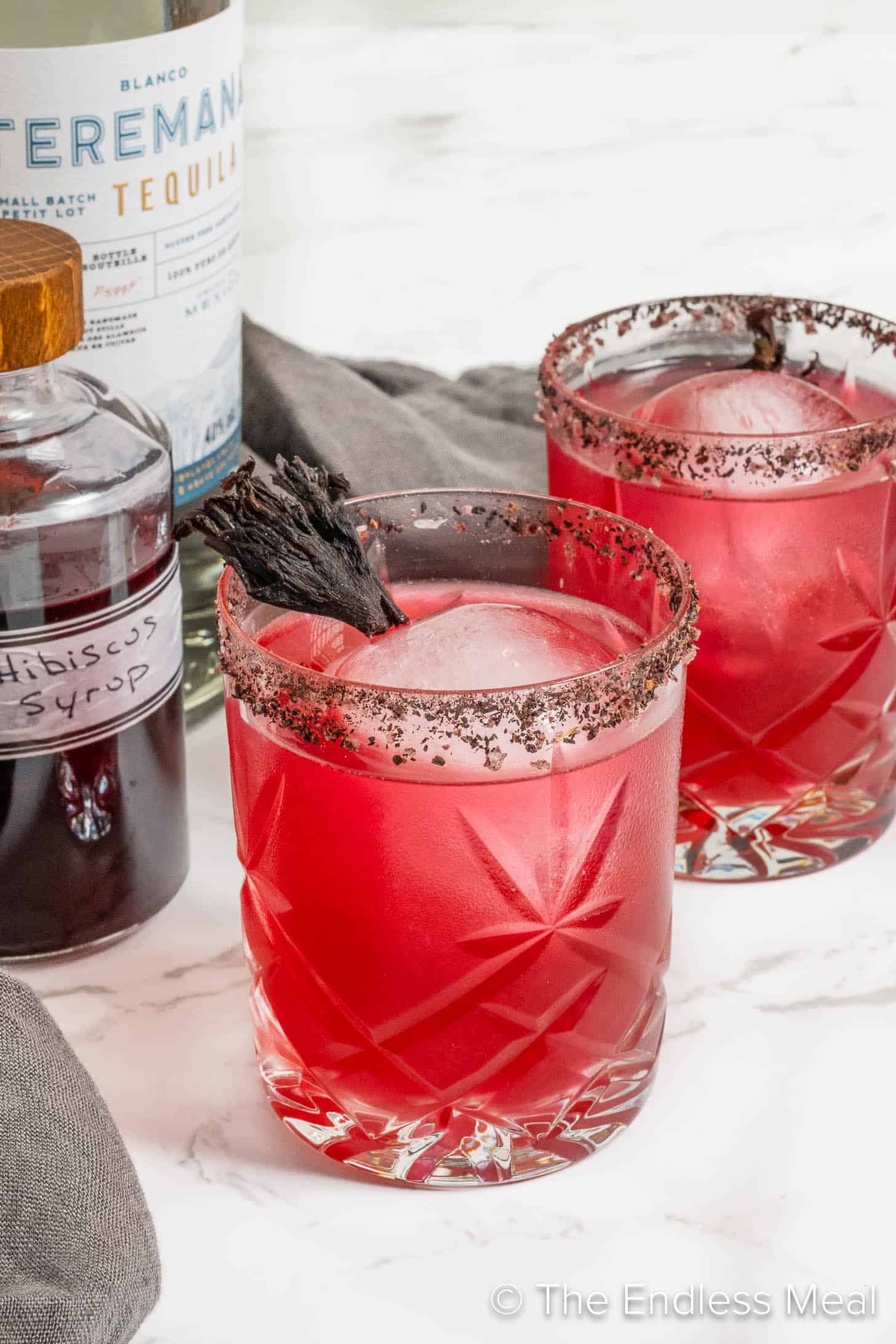Two Hibiscus Margaritas in salt rimmed glasses next to a bottle of hibiscus syrup