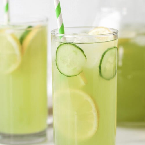 A glass of refreshing Cucumber Lemonade with ice, lemon and cucumber slices, and a green-striped straw; pitcher and another glass in the background.