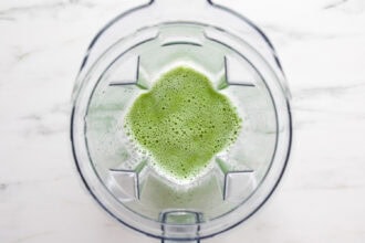Top-down view of a blender containing a green, frothy cucumber lemonade mixture on a white surface.