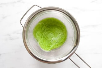 A metal strainer containing blended green cucumber lemonade is placed over a glass bowl on a white marble surface.