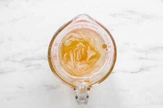 A glass measuring cup filled with a light brown Cucumber Lemonade and foam, placed on a white marble surface.