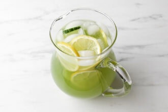 A glass pitcher filled with refreshing Cucumber Lemonade, ice cubes, cucumber slices, and lemon slices sits on a white marble surface.